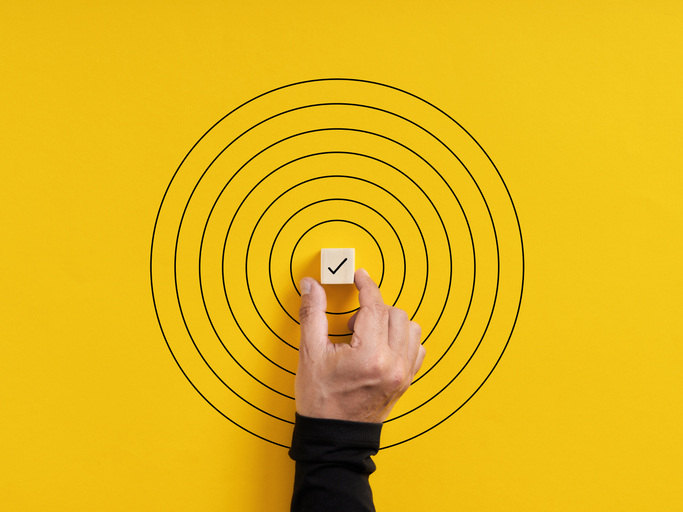 A hand places a checkmarked wooden cube in the center of a target.